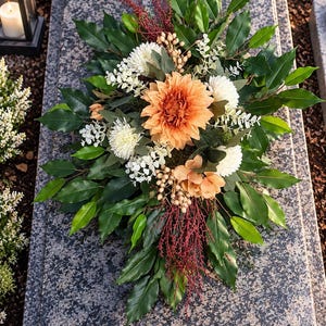 May include: A floral arrangement featuring a central peach-coloured dahlia, white chrysanthemums, and smaller peach blossoms. The bouquet is accented with dark green leaves, light green sprigs, and thin, reddish-brown decorative branches.