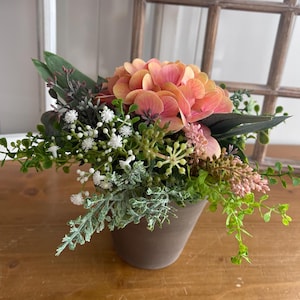 Könnte beinhalten: Blumenarrangement mit einer großen, pfirsichfarbenen Hortensienblüte, umgeben von Grün und kleinen weißen Blüten. Die Anordnung befindet sich in einem braunen Keramiktopf. Der Hintergrund umfasst einen Holzfensterrahmen.