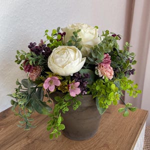 Peut inclure: Un arrangement floral dans un pot marron. Il comprend des roses blanches, des fleurs roses et violettes, et du feuillage vert. Le pot rond et texturé est posé sur une surface en bois.
