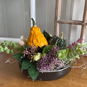 Könnte beinhalten: Eine dunkelbraune Schale mit einem dekorativen Arrangement aus einem leuchtend gelben, warzigen Kürbis, einem kleinen dunkelgrün gestreiften Kürbis, grünen eukalyptusartigen Blättern, violetten Blumenzweigen und hellen getrockneten Pflanzenteilen.