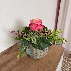 Op de afbeelding: Bloemstuk met twee roze rozen, groen blad en bessen in een grijze, getextureerde vaas met gouden accenten. Het arrangement staat op een houten oppervlak, wat een natuurlijke uitstraling creëert.