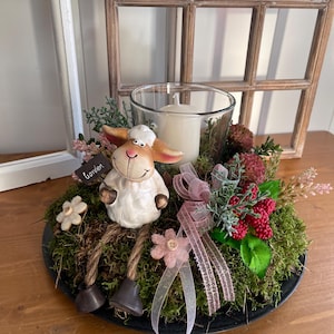 May include: A decorative centerpiece featuring a ceramic sheep figurine with a "garden" sign, surrounded by moss, flowers, and a candle in a glass holder. The arrangement includes pink ribbons and sits on a black plate.