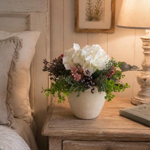 Puede incluir: Un arreglo floral en una maceta de cerámica color crema mate que presenta flores de hortensia blancas, pequeñas flores rosas, bayas de color púrpura oscuro y varias ramas de follaje verde, creando un conjunto botánico denso y redondeado.