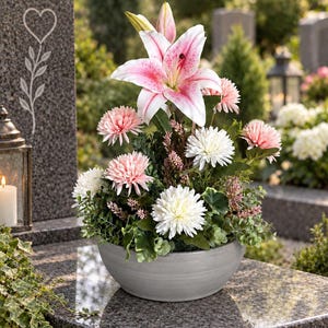 Könnte beinhalten: Ein Blumenarrangement in einer grauen Schale mit einer großen rosa-weißen Lilie, mehreren rosa und weißen chrysanthemenartigen Blüten und grünem Blattwerk. Das Arrangement steht auf einer polierten grauen Steinfläche mit Herzgravur.