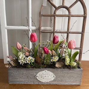 May include: A wooden rectangular planter box filled with pink tulips, white flowers, and greenery. The box has a decorative metal label that reads "Flowers & Garden". A wooden arched window frame is in the background.