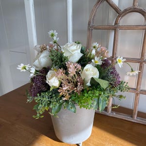 Op de afbeelding: Een bloemstuk in een grijze pot met drie kleine voetjes. Het arrangement bevat witte rozen, madeliefjes en groen. De achtergrond omvat een houten boogvenster en een wit raam.