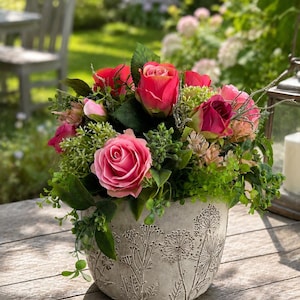 Op de afbeelding: Een bloemstuk met felroze rozen en groen blad in een ronde, lichtgrijze plantenbak met steenstructuur. De plantenbak is versierd met een reliëf van botanische patronen van wilde bloemen en paardenbloemzaadjes.