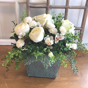 Op de afbeelding: Een bloemstuk met grote witte rozen, kleine witte bloemen en groen blad. De bloemen staan in een vierkante, getextureerde, blauwgroene metalen container. Op een houten oppervlak.