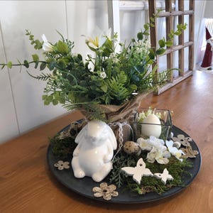 May include: A decorative Easter centerpiece on a dark gray tray. It features a white ceramic bunny, faux greenery, white flowers, a candle in a glass holder, and decorative butterflies and flowers. The arrangement is set on a wooden table.