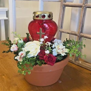 Peut inclure: Un photophore rouge en forme de cœur avec un couvercle doré et une croix, entouré de fleurs blanches, rouges et roses dans un pot en terre cuite. L'arrangement comprend de la verdure et est posé sur un fond rustique.