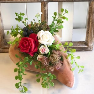 Könnte beinhalten: Dekorative Blumengesteck in einem pfirsichfarbenen, schuhförmigen Behälter. Es enthält rote und weiße Rosen, kleine rosa Blüten und grüne Blätter. Das Arrangement ist für die Inneneinrichtung gedacht.