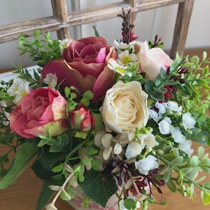 Könnte beinhalten: Ein Blumenarrangement mit rosa und cremefarbenen Rosen, kleinen weißen Blüten und grünem Laub. Die Blumen sind in einer gemusterten Vase angeordnet. Im Hintergrund befindet sich ein Holzfensterrahmen.