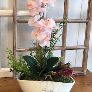 Op de afbeelding: Decoratief bloemstuk met lichtroze orchideeën, groene bladeren en rode bessen in een witte keramische kom. De kom heeft een gebogen vorm en is gemarkeerd met "Cerastyle". De opstelling staat voor een houten raamkozijn.