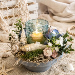 May include: A grey bowl centrepiece featuring a light blue glass candle holder with white fish illustrations, a gold fish figurine, assorted seashells, green moss, and small white and blue flowers, with a lit yellow candle inside the glass.