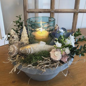 Könnte beinhalten: Dekoratives Tischarrangement mit einer brennenden Kerze in einem tealfarbenen Glasbehälter mit Fischmotiven. Die Anordnung umfasst Muscheln, eine goldene Fischfigur, Grünzeug und weiße Blumen, alles in einer grauen Schale.