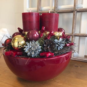 Könnte beinhalten: Festlicher Mittelpunkt in einer glänzend roten Schale mit zwei burgunderfarbenen Kerzen, goldenen und roten Ornamenten, Tannenzapfen und dekorativen Akzenten. Geeignet für die Weihnachtsdekoration.