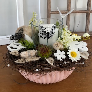 Könnte beinhalten: Eine rosa geriffelte Schale gefüllt mit braunen Zweigen, einer weißen Kerze im Glas, einem schwarz-weißen Schmetterlingsornament, einer grünen Sukkulente, kleinen Muscheln, weißen Kunstblumen und einer Schnur mit kleinen weißen Perlen.