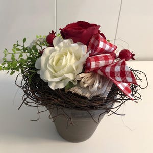 Puede incluir: Arreglo floral decorativo en una maceta gris. El arreglo presenta una cinta de cuadros rojos y blancos, rosas rojas y blancas, follaje verde y pequeñas bayas rojas, todo anidado en un nido de ramitas tejidas.
