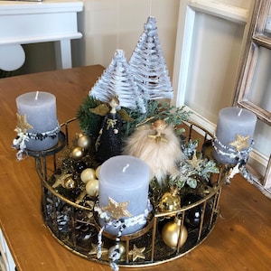 Op de afbeelding: Een decoratief kerststuk met vier grijze kaarsen, gouden sterren en engelenbeeldjes. Het middelpunt staat in een ronde, goudkleurige metalen lijst, versierd met ornamenten en miniatuur kerstbomen.