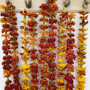 May include: Five long garlands of vibrant orange and deep red marigold flowers hang vertically from a light-colored wooden wall rack. The rack features five brass-toned animal-shaped hooks, including a rabbit, fox, deer, owl, and squirrel.