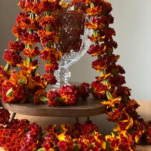 May include: A clear, faceted glass pedestal vase draped with a long garland of vibrant orange and deep red marigold flowers. The floral garland hangs over the sides of the glass vessel, creating a layered display of warm-toned, ruffled blossoms.