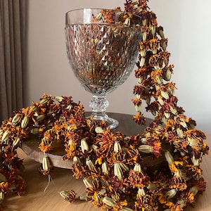 May include: A clear glass goblet with a diamond-cut pattern holds a long, draped garland of dried marigold flowers. The garland features deep orange and dark maroon petals with light green calyxes, cascading from the rim of the glass vessel.
