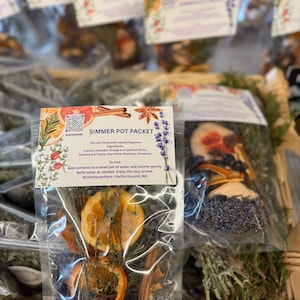 Puede incluir: Paquetes transparentes llenos de hierbas secas, especias y rodajas de cítricos, etiquetados como "Simmer Pot Packet". Los ingredientes incluyen lavanda, naranja, pomelo, romero, tomillo, anís estrellado, escaramujos y canela. Los paquetes se exhiben en una cesta de mimbre.