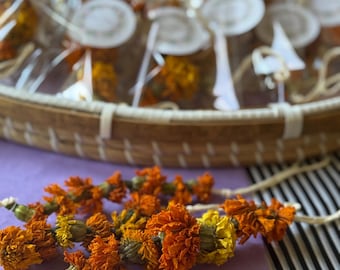 May include: A garland of dried orange and yellow marigold flowers strung on a thin white cord. The flowers are densely packed along the string, showcasing natural textures and vibrant autumn colors. Additional packaged floral items are visible.