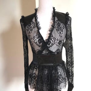 Può includere: Top in pizzo nero con maniche lunghe e orlo a peplo. Il top presenta uno scollo a V con finiture a balze e una cintura nera in similpelle. L'indumento è esposto su un manichino.