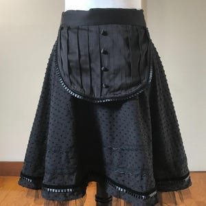 May include: Black skirt with a unique design. The skirt features a pleated panel with buttons, a dotted pattern, and decorative trim. A layer of black tulle adds a delicate touch to the hemline. The skirt is displayed on a black mannequin.