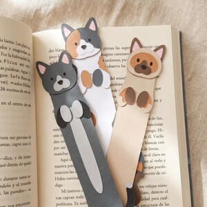 Puede incluir: Tres marcapáginas con forma de gato dentro de un libro abierto. Un marcapáginas es gris oscuro con el pecho blanco, otro es blanco con manchas naranjas y grises, y otro es marrón claro con la cara y las orejas de color marrón oscuro.