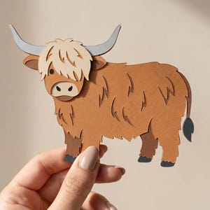 Puede incluir: Un recorte de papel en capas de una vaca Highland con cara de color tostado claro, cuerpo marrón peludo y cuernos largos de color gris. La vaca tiene una nariz de color marrón oscuro, ojos pequeños negros y pezuñas de color gris oscuro.