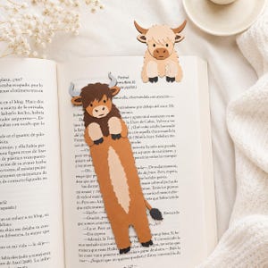 May include: Two paper bookmarks shaped like Highland cattle. One is a long, brown, shaggy-haired cow with grey horns and a light tan belly stripe. The other is a smaller, cream-colored cow head with brown horns, both resting on an open book.