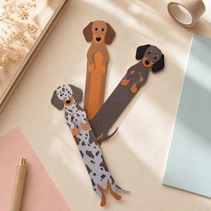 May include: Three dachshund dog bookmarks in various colors. One is brown, one is gray with white spots, and the other is dark brown. Each bookmark has a long body and is made of paper.