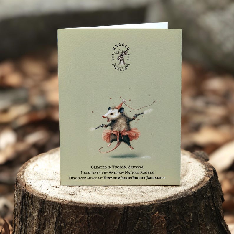 K&ouml;nnte beinhalten: Gru&szlig;karte mit einer skurrilen Illustration eines Opossums mit Tutu und Partyhut, das zwei Pistolen h&auml;lt. Die Karte enth&auml;lt den Text "RUGGED JACKALOPE" und "CREATED IN TUCSON, ARIZONA". Die Illustration stammt von Andrew Nathan Rogers.