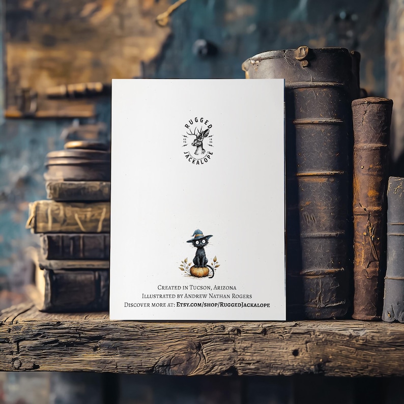 Pu&ograve; includere: Un biglietto di auguri bianco con un gatto nero con un cappello su una zucca. Il biglietto ha il testo "RUGGED JACKALOPE" e "CREATED IN TUCSON, ARIZONA". Il biglietto &egrave; su una mensola di legno con vecchi libri.