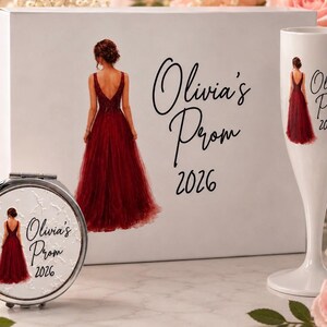 May include: A white rectangular gift box, a round silver compact mirror, and a white champagne flute. Each item features a graphic of a person in a deep red gown and the black text Olivia's Prom 2026. The items are arranged together.