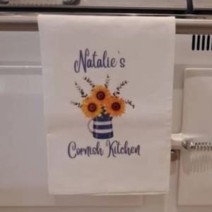 May include: White kitchen towel with the text "Natalie's Cornish Kitchen" in dark blue script. The towel features a watercolour illustration of sunflowers in a blue and white striped jug.