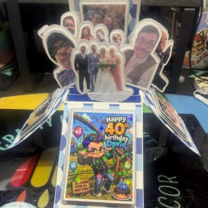 May include: A handmade 40th birthday card with a pop-up design. The card includes photos of people and a central image with the text "Happy 40th birthday David". The card has a blue and white colour scheme with glitter accents.