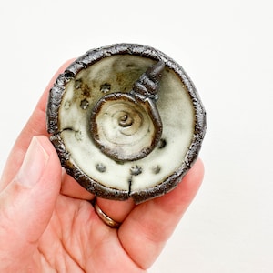 May include: Handmade ceramic dish with a spiral design in the center. The dish has a light green interior with dark brown edges and a textured surface. The dish is held in a hand against a white background.