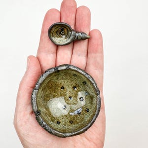 May include: Two small, handmade ceramic bowls. The larger bowl is round with a textured, dark rim and a speckled, olive-green interior with star-shaped accents. The smaller bowl is snail-shaped, with a similar color palette.