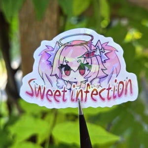 Pode incluir: Um autocolante hologr&aacute;fico com uma personagem de estilo anime com cabelo rosa, um halo e uma seringa. O autocolante tem as palavras "Sweet Infection" em escrita rosa. O fundo &eacute; folhagem verde desfocada.