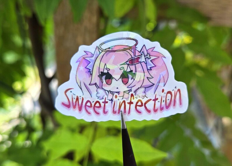 Pode incluir: Um autocolante hologr&aacute;fico com uma personagem de estilo anime com cabelo rosa, um halo e uma seringa. O autocolante tem as palavras "Sweet Infection" em escrita rosa. O fundo &eacute; folhagem verde desfocada.