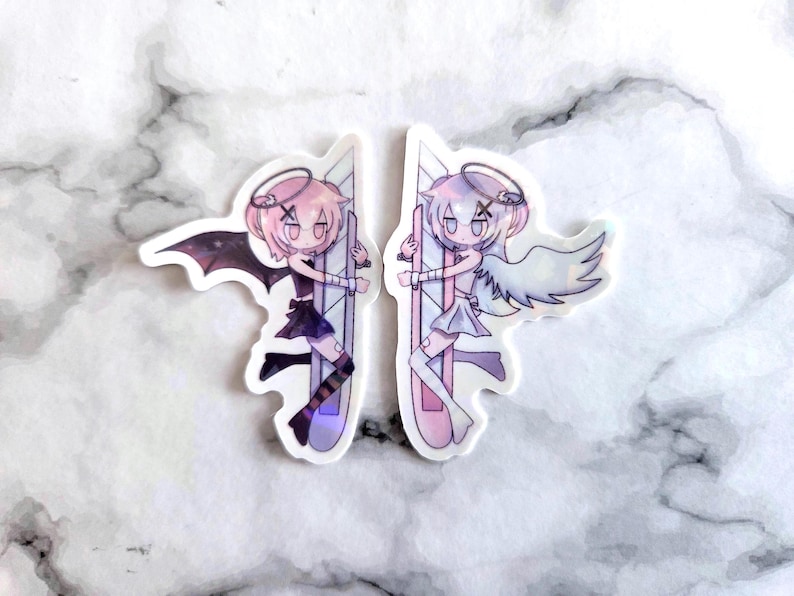 May include: Two stickers featuring anime-style characters, one a devil with black wings and the other an angel with white wings. Both characters have pink hair, halos, and are holding a stylized sword. The stickers are on a white marble background.