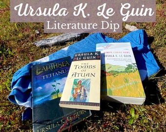 Puede incluir: Varios libros de Ursula K. Le Guin, incluyendo "Tehanu", "The Tombs of Atuan" y "Always Coming Home", se muestran sobre una tela azul. La imagen también presenta el texto "Ursula K. Le Guin Literature Dip."