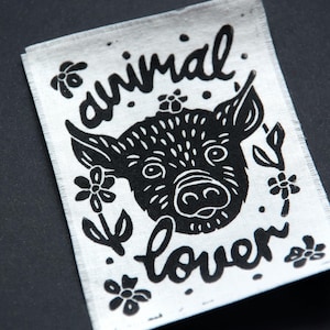 Könnte beinhalten: Ein weißes Stoffpatch mit einer schwarzen Illustration eines Schweinegesichts, umgeben von floralen Akzenten und den Worten "animal lover". Das Schwein hat große Ohren und eine detaillierte Schnauze. Der Text ist handschriftlich.