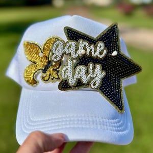 May include: White trucker hat with a curved brim, featuring a gold fleur-de-lis and a black star, both embellished with sequins. The words "game day" are written in white sequins. The hat is set against a blurred green background.