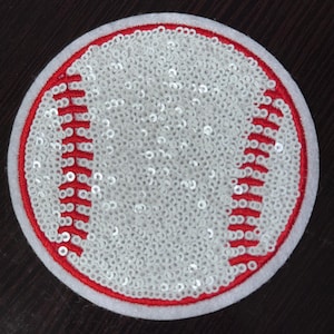 Puede incluir: Un parche de béisbol blanco con costuras rojas, hecho de lentejuelas. El parche es redondo y tiene un borde blanco.
