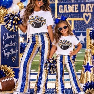 Puede incluir: Dos camisetas blancas con un gráfico de un balón de fútbol americano y pompones, combinadas con pantalones de campana a juego con rayas de lentejuelas azules, doradas y plateadas. Los conjuntos incluyen zapatillas blancas con estrellas azules.