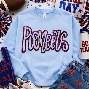 May include: A light blue crewneck sweatshirt featuring the word Pioneers in white and red sequined script lettering across the chest. The garment has long sleeves, a ribbed neckline, and a standard fit, showcasing a textured, sparkling finish.